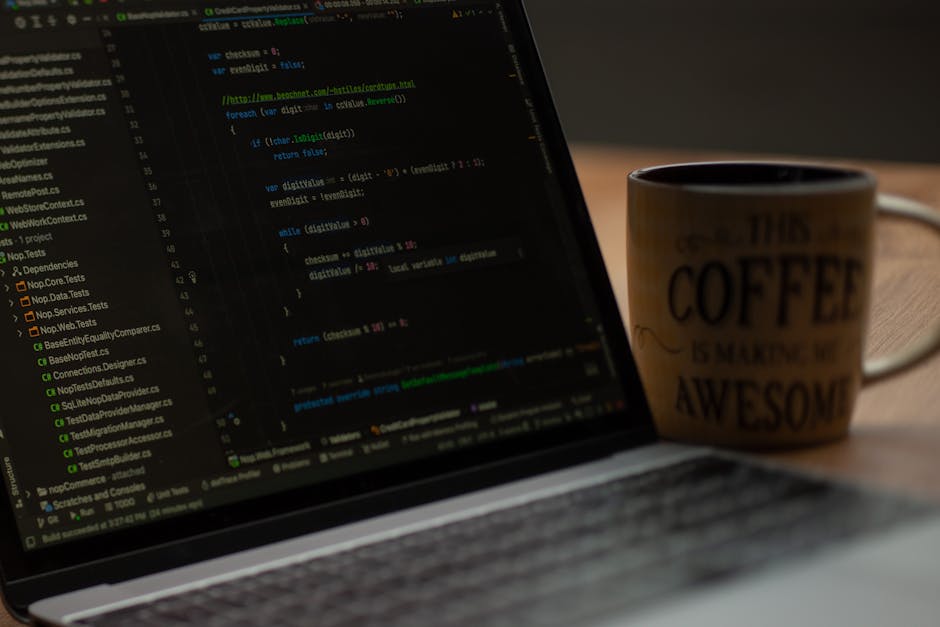A close-up view of a laptop displaying lines of code on a dark background, with various colourful syntax highlighting. Sitting beside the laptop is a ceramic coffee mug with the words 'This coffee is 