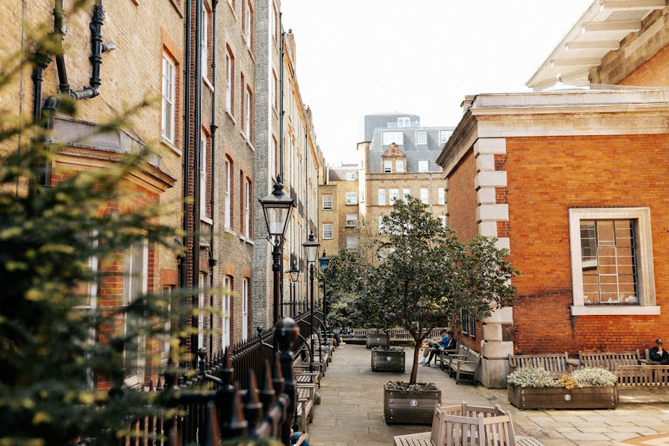 A serene urban courtyard surrounded by historic brick and stone buildings with classic window designs, featuring a paved pathway lined with black wrought-iron lamp posts and a variety of landscaping e